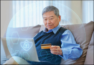 An older man on a sofa holds a bank card while using a laptop